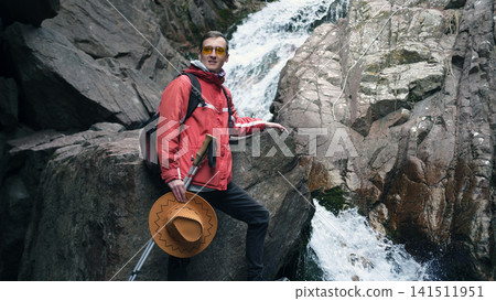 A young student travels around his country. A guy in a red jacket and yellow glasses stands against the backdrop of a waterfall A young student travels around his country. A guy in a red jacket and yellow glasses stands against the backdrop of a waterfall 141511951