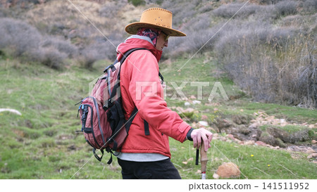 A lonely wanderer travels through the mountains. A young guy in a cowboy hat, backpack, trekking poles and yellow glasses walks through a green field A lonely wanderer travels through the mountains. A young guy in a cowboy hat, backpack, trekking poles and yellow glasses walks through a green field 141511952
