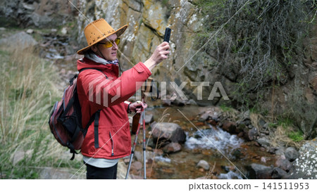A young guy in a cowboy hat and a red jacket is photographing a river near a rocky mountain. A lone traveler walks through the expanses of the canyon 141511953
