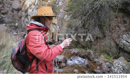 A funny tourist in a cowboy hat and yellow glasses looks at his phone while hiking along a mountain river. Walking alone A funny tourist in a cowboy hat and yellow glasses looks at his phone while hiking along a mountain river. Walking alone 141511954