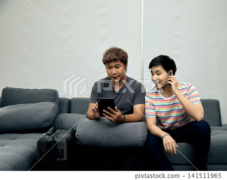 Two men sitting on a sofa using a tablet and phone beside luggage, planning travel, booking accommodation, communicating, browsing, friendship, technology, and departure preparation Two men sitting on a sofa using a tablet and phone beside luggage, planning travel, booking accommodation, communicating, browsing, friendship, technology, and departure preparation 141511965