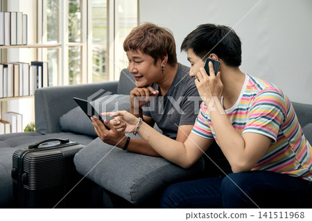 Two men sitting on a sofa using a tablet and phone beside luggage, planning travel, booking a trip, discussing details, communicating, browsing, technology, friendship, and vacation preparation Two men sitting on a sofa using a tablet and phone beside luggage, planning travel, booking a trip, discussing details, communicating, browsing, technology, friendship, and vacation preparation 141511968