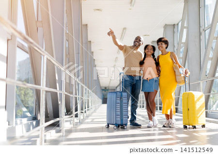 Black father pointing ahead while standing with wife and daughter in airport 141512549