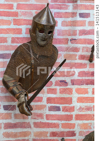 Medieval knight in chainmail stands against brick wall. Helmet with pointed top covers his face. He grips a sword with both hands. Brick wall behind him is textured and red 141513143