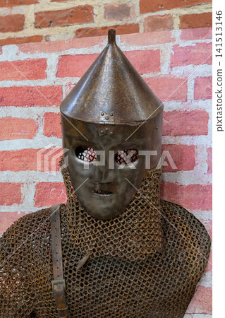 Ancient metal helmet with chainmail armor. Red brick wall background. Helmet has eye slits and pointed top. Armor shows weathered, historical texture. Suitable for medieval or fantasy themes Ancient metal helmet with chainmail armor. Red brick wall background. Helmet has eye slits and pointed top. Armor shows weathered, historical texture. Suitable for medieval or fantasy themes 141513146