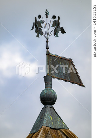 Weather vane atop wooden roof with bird figures. Birds sculpted in green metal, wings spread. Weather vane shows signs of age and exposure. Cloudy sky forms soft backdrop behind Weather vane atop wooden roof with bird figures. Birds sculpted in green metal, wings spread. Weather vane shows signs of age and exposure. Cloudy sky forms soft backdrop behind 141513155