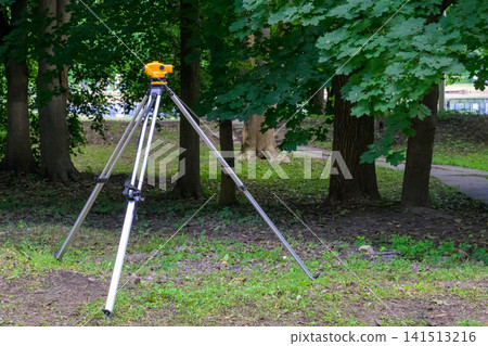 Yellow surveyor mounted on a silver tripod outdoors. Trees with green leaves surround the setup. Tripod stands on grassy ground near a dirt path. Natural daylight illuminates the scene clearly Yellow surveyor mounted on a silver tripod outdoors. Trees with green leaves surround the setup. Tripod stands on grassy ground near a dirt path. Natural daylight illuminates the scene clearly 141513216