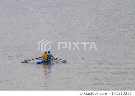 Two people row a small boat on calm water. They use oars to propel the vessel forward. The water is gentle with subtle ripples. The rowers wear bright tops for visibility. Their motion suggests steady Two people row a small boat on calm water. They use oars to propel the vessel forward. The water is gentle with subtle ripples. The rowers wear bright tops for visibility. Their motion suggests steady 141513285