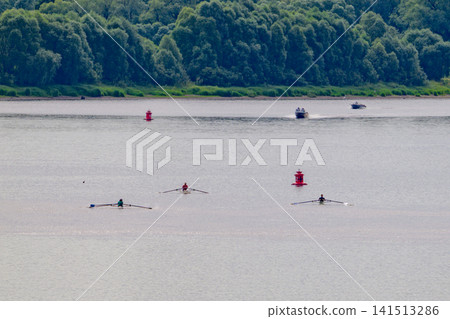 Rowers glide across calm lake waters. Red buoys mark racing course in distance. Trees line shoreline behind wide expanse. Boat trails wake gently behind rowers. Clear day enhances natural scenery Rowers glide across calm lake waters. Red buoys mark racing course in distance. Trees line shoreline behind wide expanse. Boat trails wake gently behind rowers. Clear day enhances natural scenery 141513286