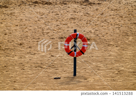 Orange lifebuoy stands on sandy beach. Black post holds it upright. Deserted sand stretches wide around. Minimalist beach safety setup. No animals or plants present. Sole focus: life-saving device Orange lifebuoy stands on sandy beach. Black post holds it upright. Deserted sand stretches wide around. Minimalist beach safety setup. No animals or plants present. Sole focus: life-saving device 141513288