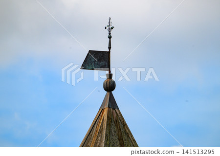 Weather vane atop a conical roof under a partly cloudy sky. Metal weather vane shows signs of age and exposure. Crafted with decorative finial and directional arrow Weather vane atop a conical roof under a partly cloudy sky. Metal weather vane shows signs of age and exposure. Crafted with decorative finial and directional arrow 141513295