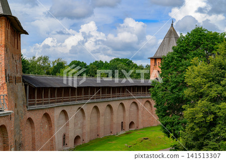 Ancient stone fortress with arched walkway. Weathered walls surround green grassy area. Tower rises above dense tree canopy. Clouds drift across bright blue sky Ancient stone fortress with arched walkway. Weathered walls surround green grassy area. Tower rises above dense tree canopy. Clouds drift across bright blue sky 141513307