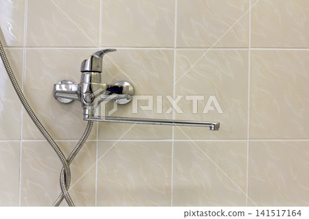 Water tap, chrome stainless metal faucet connected to shower stall tiled wall, mixer cold hot water on blurred bathroom copy space background. Modern home interior. Water tap, chrome stainless metal faucet connected to shower stall tiled wall, mixer cold hot water on blurred bathroom copy space background. Modern home interior. 141517164