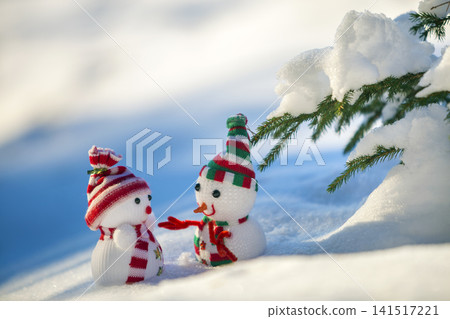 Two small funny toys baby snowman in knitted hats and scarves in deep snow outdoors near pine tree branch. Happy New Year and Merry Christmas greeting card. Two small funny toys baby snowman in knitted hats and scarves in deep snow outdoors near pine tree branch. Happy New Year and Merry Christmas greeting card. 141517221