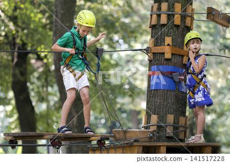 Two children, boy and girl in protective harness and safety helmets at climbing activity on rope way. Two children, boy and girl in protective harness and safety helmets at climbing activity on rope way. 141517228