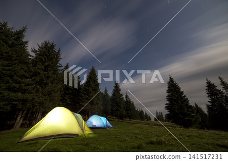 Two brightly lit tourist tents on green grassy forest clearing among tall pine trees on clear dark blue starry sky background. Tourism, night camping in summer mountains, beauty of nature concept. Two brightly lit tourist tents on green grassy forest clearing among tall pine trees on clear dark blue starry sky background. Tourism, night camping in summer mountains, beauty of nature concept. 141517231