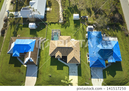 Top view of leaking house roof covered with protective tarp sheets against rain water leaks until replacement of asphalt shingles. Damage of building rooftop as aftermath of hurricane Ian in Florida Top view of leaking house roof covered with protective tarp sheets against rain water leaks until replacement of asphalt shingles. Damage of building rooftop as aftermath of hurricane Ian in Florida 141517250