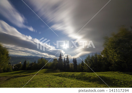 Summer night mountain panorama. Green trees on grassy mountain clearing on cloudy evening sky copy space background, woody hills on horizon, bright dwelling and road lights in valley. Summer night mountain panorama. Green trees on grassy mountain clearing on cloudy evening sky copy space background, woody hills on horizon, bright dwelling and road lights in valley. 141517294