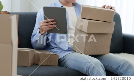 Young Asian woman organizing cardboard parcels while using digital tablet on sofa during house move Young Asian woman organizing cardboard parcels while using digital tablet on sofa during house move 141520010