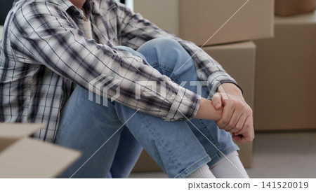 Asian man sitting on floor holding arm during moving break surrounded by cardboard boxes in new apartment 141520019