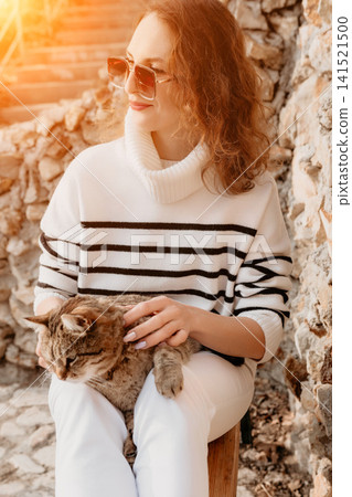 Woman cat pet sitting outside in striped sweater and sunglasses petting a tabby on her lap in warm golden sunlight 141521500