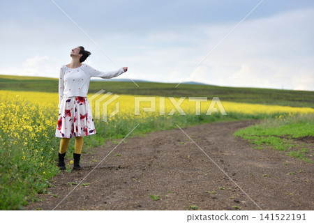 Woman hitchhiking on a dirt road next to a yellow canola flower field 141522191