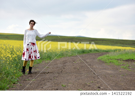Woman hitchhiking on a dirt road next to a yellow canola flower field Woman hitchhiking on a dirt road next to a yellow canola flower field 141522192