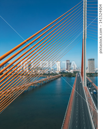 aerial drone view of suspension cable bridge over Han river in Danang, geometric cable-stayed bridge architecture with modern city skyline and roadway, Da Nang Vietnam aerial drone view of suspension cable bridge over Han river in Danang, geometric cable-stayed bridge architecture with modern city skyline and roadway, Da Nang Vietnam 141524904