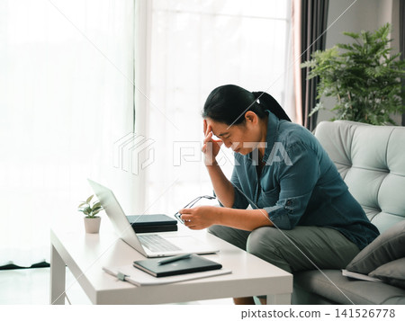 Stressed woman sitting near laptop at home, holding eyeglasses and touching her forehead, showing headache, digital eye strain, work fatigue, and remote work burnout. Stressed woman sitting near laptop at home, holding eyeglasses and touching her forehead, showing headache, digital eye strain, work fatigue, and remote work burnout. 141526778