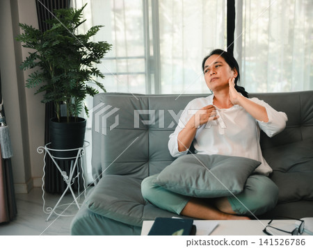 Middle-aged woman sitting on sofa and touching her neck, showing neck pain, body discomfort, office syndrome, fatigue, stress, and health care concept at home. Middle-aged woman sitting on sofa and touching her neck, showing neck pain, body discomfort, office syndrome, fatigue, stress, and health care concept at home. 141526786