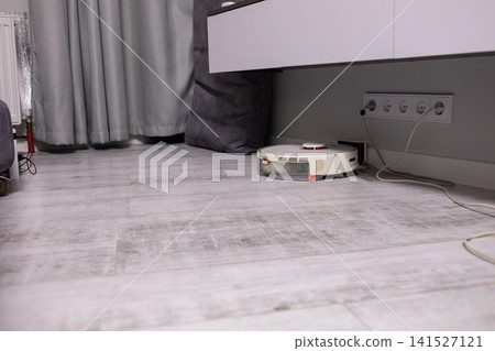 Low angle view of a robot vacuum cleaner moving across a light kitchen floor beneath modern cabinets, illustrating automated housekeeping, smart cleaning and daily home care tasks Low angle view of a robot vacuum cleaner moving across a light kitchen floor beneath modern cabinets, illustrating automated housekeeping, smart cleaning and daily home care tasks 141527121