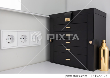 Black wooden jewelry organizer with small drawers stands on a light shelf beside decorative figurines and wall outlets, showing elegant home storage and modern interior decor view Black wooden jewelry organizer with small drawers stands on a light shelf beside decorative figurines and wall outlets, showing elegant home storage and modern interior decor view 141527124
