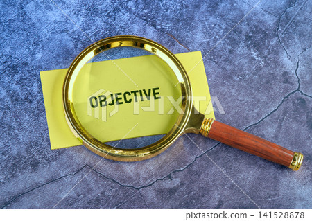 Gold magnifying glass rests on a bright yellow card to emphasize the word Objective during a business strategy review on a dark table surface Gold magnifying glass rests on a bright yellow card to emphasize the word Objective during a business strategy review on a dark table surface 141528878