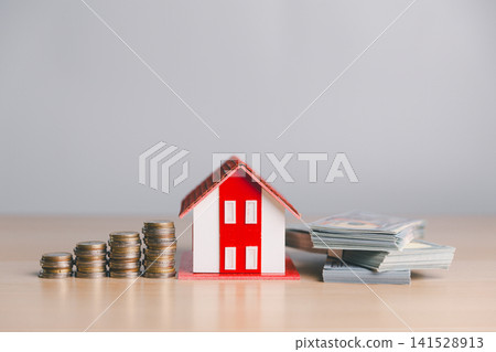 Miniature red house with coin stacks and cash bundles on wooden table, representing home loan, property investment, mortgage payment, savings, and real estate value. Miniature red house with coin stacks and cash bundles on wooden table, representing home loan, property investment, mortgage payment, savings, and real estate value. 141528913