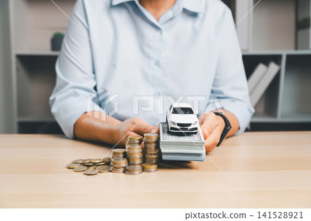 White toy car placed on a stack of cash with coins on desk, symbolizing car purchase cost, auto loan, vehicle finance, cash payment, down payment, and automobile ownership. White toy car placed on a stack of cash with coins on desk, symbolizing car purchase cost, auto loan, vehicle finance, cash payment, down payment, and automobile ownership. 141528921