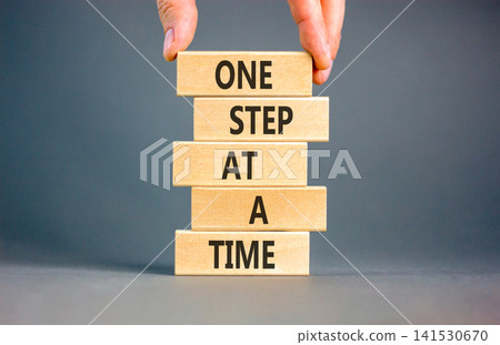 One step at a time symbol. Concept words One step at a time on beautiful wooden blocks. Beautiful grey background. Businessman hand. Business and one step at a time concept. Copy space. One step at a time symbol. Concept words One step at a time on beautiful wooden blocks. Beautiful grey background. Businessman hand. Business and one step at a time concept. Copy space. 141530670