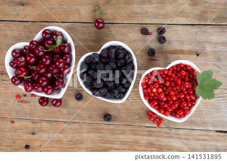 Three heart shaped white bowls filled with cherries blackberries and redcurrants on rustic wooden tabletop, viewed from above with scattered berries, fresh leaves and vibrant color 141531895