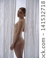 Elegant nude woman looking back framed by soft linen curtains 141532718