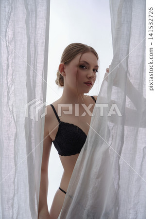 Elegant woman in black lingerie looking from behind sheer white curtain Elegant woman in black lingerie looking from behind sheer white curtain 141532726