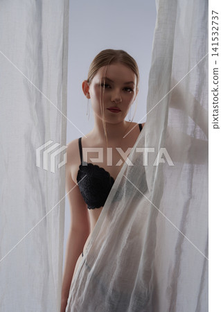 Elegant woman in black lingerie looking from behind sheer white curtain 141532737