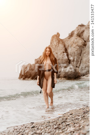 Woman beach shoreline walking in swimsuit and robe on pebble beach with rocky cliffs at sunset Woman beach shoreline walking in swimsuit and robe on pebble beach with rocky cliffs at sunset 141533477