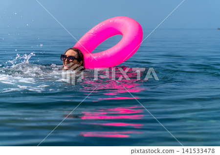 Woman swimming cheerfully with bright pink inflatable ring in deep blue summer ocean water, vacation freedom concept Woman swimming cheerfully with bright pink inflatable ring in deep blue summer ocean water, vacation freedom concept 141533478