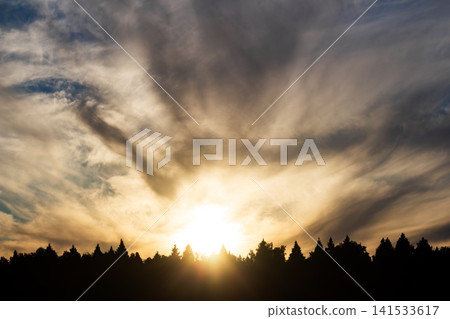 Dramatic sunset sky with intense glowing sun rays piercing through textured clouds above a dark forest silhouette, capturing a serene and atmospheric evening landscape in nature 141533617