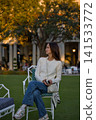 Woman In White Sweater Sitting On A Garden Chair Under A Tree At Dusk 141533772