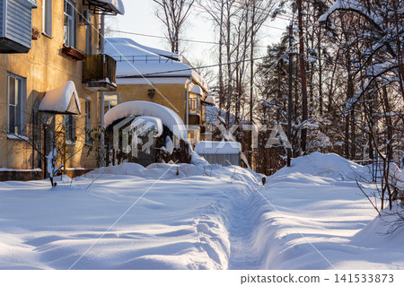 Sunlit residential house covered in deep fluffy snow with a narrow path winding through the yard against a backdrop of bare winter trees in the quiet frosty afternoon Sunlit residential house covered in deep fluffy snow with a narrow path winding through the yard against a backdrop of bare winter trees in the quiet frosty afternoon 141533873