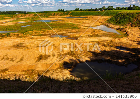 Wide landscape view of an open pit sand mine with textured ground and small water pools under a bright blue sky, revealing rugged terrain and industrial remnants in nature Wide landscape view of an open pit sand mine with textured ground and small water pools under a bright blue sky, revealing rugged terrain and industrial remnants in nature 141533900