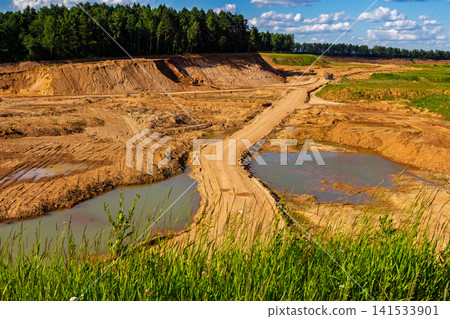 Wide panoramic shot of an active sand quarry with heavy excavation machinery, steep sandy slopes, dirt roads, and small rain ponds under a bright blue summer sky with light clouds Wide panoramic shot of an active sand quarry with heavy excavation machinery, steep sandy slopes, dirt roads, and small rain ponds under a bright blue summer sky with light clouds 141533901