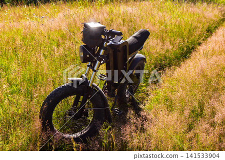 Modern black electric bicycle standing in the middle of a golden grassy field under bright warm summer sunlight, stylish fat tire e-bike ready for off-road adventure in nature 141533904