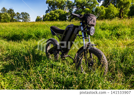 Modern matte black electric bike parked in thick green grass on a summer day, featuring sturdy fat tires and a small handlebar bag for adventurous off-road eco-friendly travel Modern matte black electric bike parked in thick green grass on a summer day, featuring sturdy fat tires and a small handlebar bag for adventurous off-road eco-friendly travel 141533907