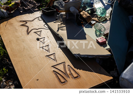 Large wooden panel with etched palm tree design lying amidst a chaotic pile of discarded household waste, glass jars, and plastic debris at a rural disposal site Large wooden panel with etched palm tree design lying amidst a chaotic pile of discarded household waste, glass jars, and plastic debris at a rural disposal site 141533910